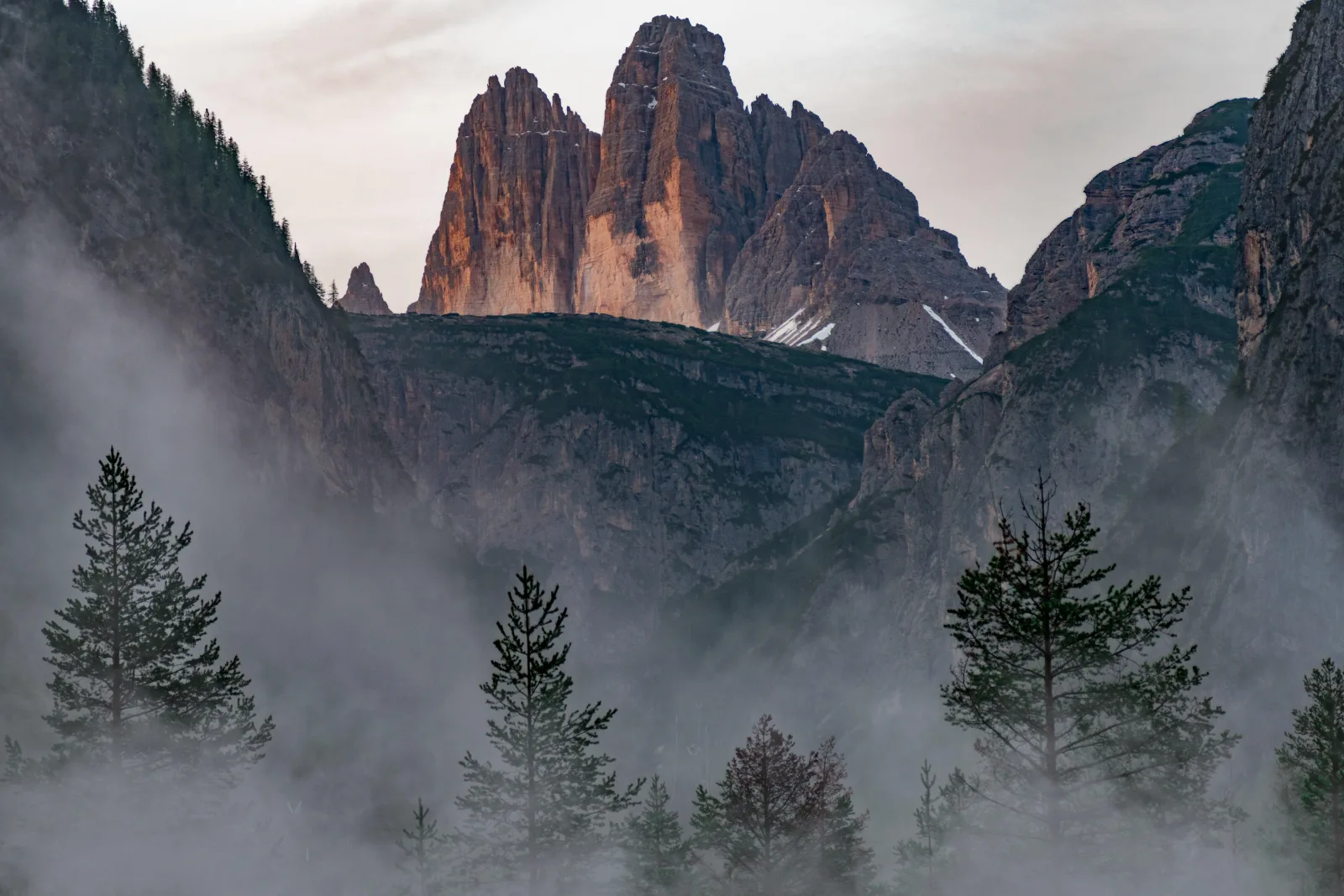 The Tre Cime di Lavaredo rising above mist and dark conifers in the valley below