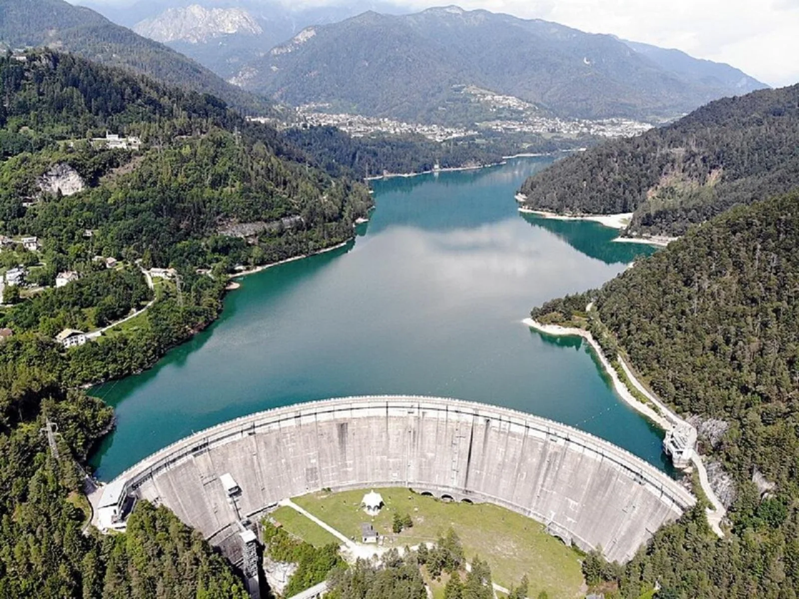 Aerial view of the curved concrete dam at the head of a green Dolomites reservoir